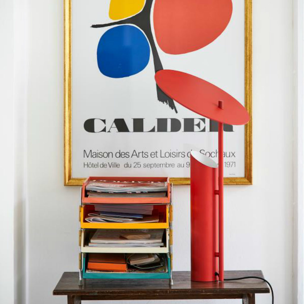 The red Reflect Table Lamp in front of an Alexander Calder poster