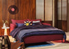 A modern upholstered bed in a warm wood-paneled room, dressed with deep purple bedding and a blue throw blanket, with decorative pillows including a yellow star accent.