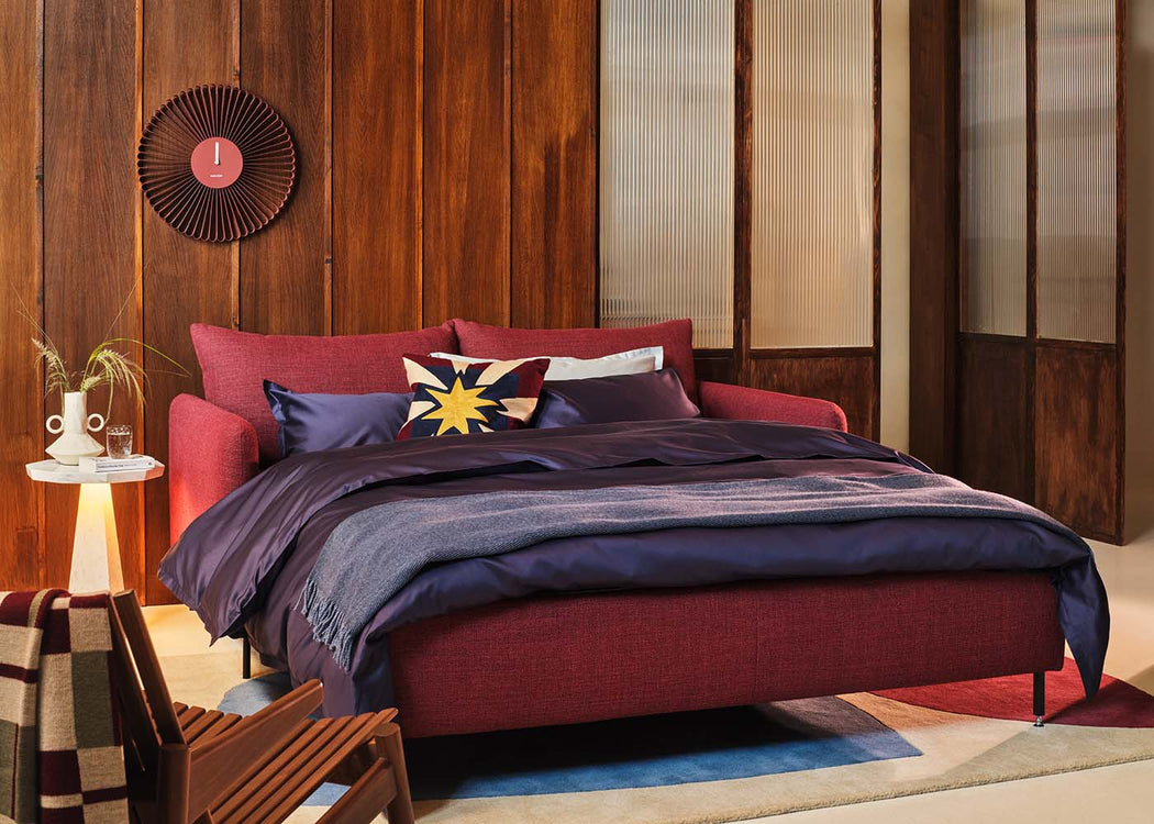A modern upholstered bed in a warm wood-paneled room, dressed with deep purple bedding and a blue throw blanket, with decorative pillows including a yellow star accent.