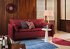 A red upholstered loveseat sofa in a mid-century modern living room setting with wood-paneled walls, a blue rug, a white side table with a lamp, and patterned throw pillows.