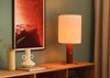 A lit table lamp with a warm cream fabric shade and a reddish-brown cylindrical base sits on a wooden console table in a styled room setting with a framed red artwork, a ceramic vase, and a curtain.