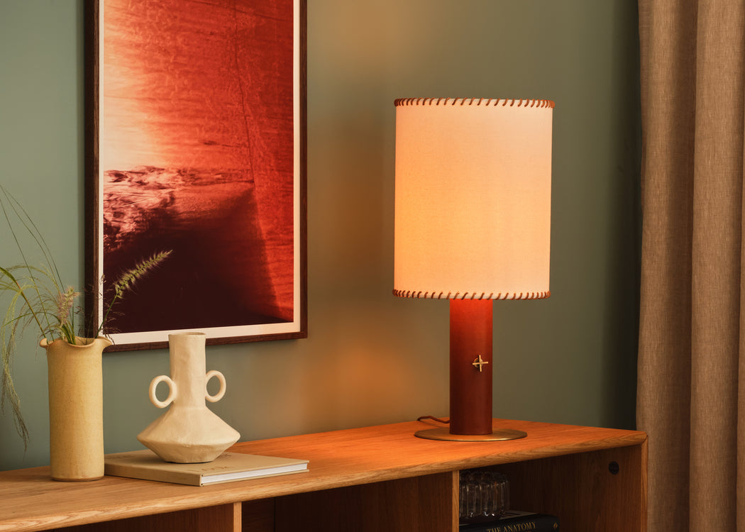 A lit table lamp with a warm cream fabric shade and a reddish-brown cylindrical base sits on a wooden console table in a styled room setting with a framed red artwork, a ceramic vase, and a curtain.