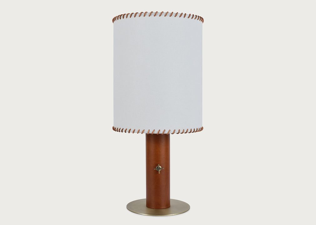 A table lamp with a white cylindrical fabric shade featuring brown leather trim at the top and bottom, mounted on a brown leather base with a round brass-colored metal foot ring, set against a solid light gray background.