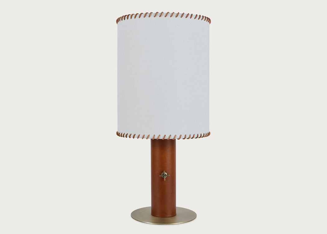 A table lamp with a white cylindrical fabric shade featuring brown leather trim at the top and bottom, mounted on a brown leather base with a round brass-colored metal foot ring, set against a solid light gray background.