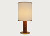 A table lamp with a warm beige fabric drum shade featuring brown leather trim, a cylindrical brown wooden base with a small gold star accent, and a round gold metal foot ring, set against a solid light gray background.