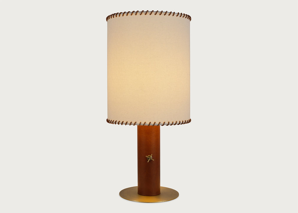 A table lamp with a warm beige fabric drum shade featuring brown leather trim, a cylindrical brown wooden base with a small gold star accent, and a round gold metal foot ring, set against a solid light gray background.