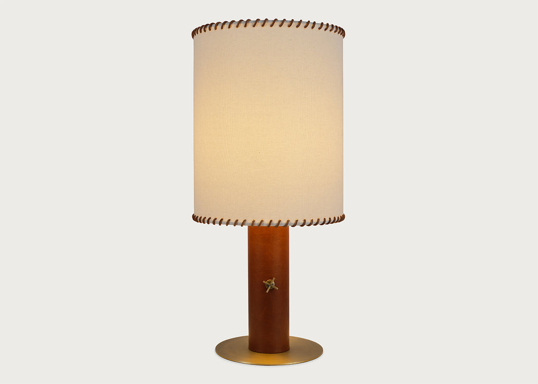 A table lamp with a warm beige fabric drum shade featuring brown leather trim, a cylindrical brown wooden base with a small gold star accent, and a round gold metal foot ring, set against a solid light gray background.