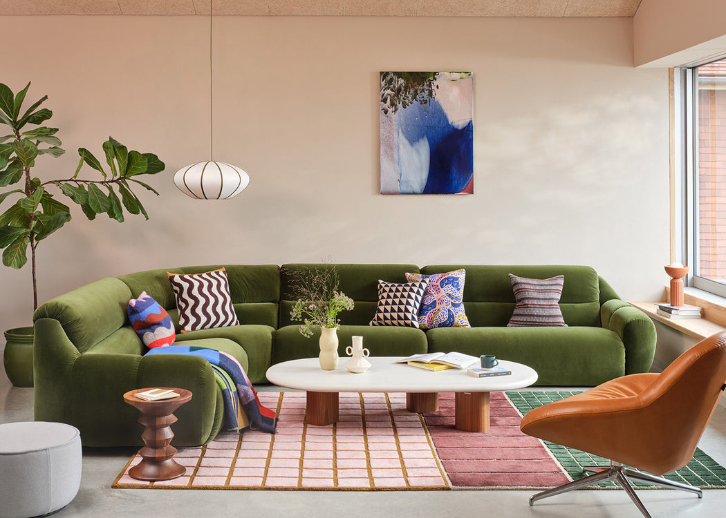 A styled living room featuring a prominent olive green sectional sofa decorated with patterned throw pillows, set against a beige wall with a blue and white abstract painting. A white oval coffee table, a pink and green striped rug, and a modern orange leather chair complete the scene.