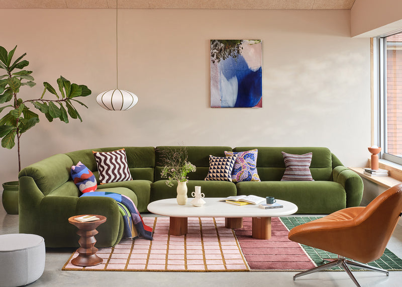 A styled living room featuring a prominent olive green sectional sofa decorated with patterned throw pillows, set against a beige wall with a blue and white abstract painting. A white oval coffee table, a pink and green striped rug, and a modern orange leather chair complete the scene.