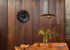 A modern black sunburst wall clock hangs on a dark wood-paneled wall above a wooden dining table with a vase of greenery and two amber glasses. A black pendant light with a warm gold interior is suspended nearby.