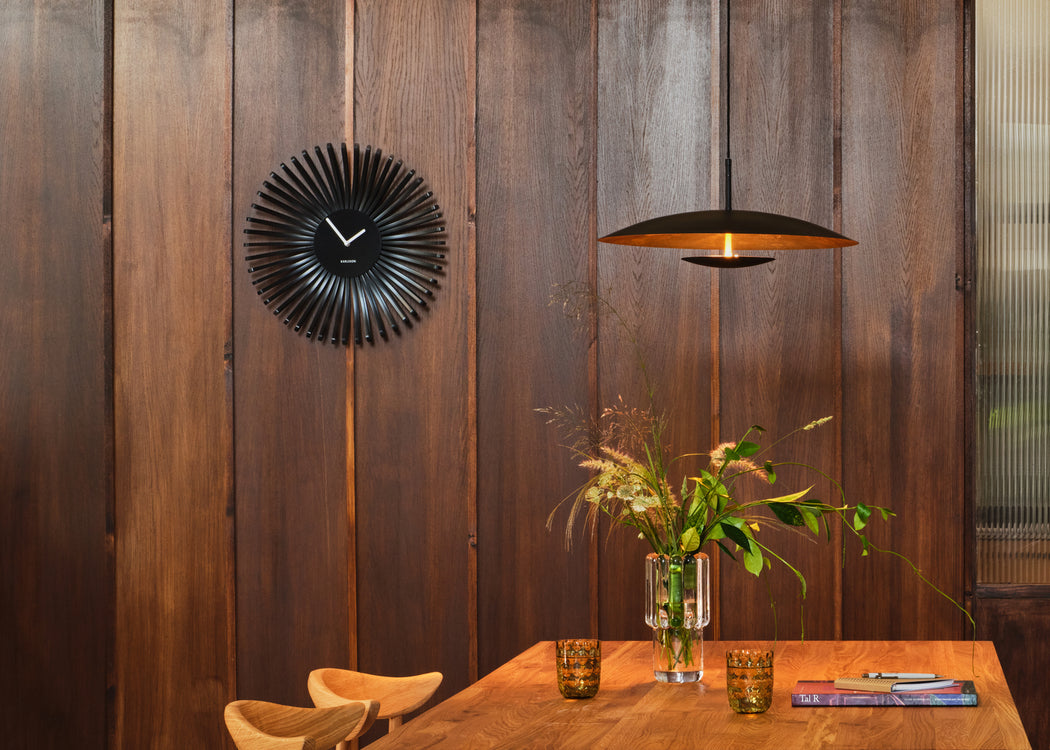 A modern black sunburst wall clock hangs on a dark wood-paneled wall above a wooden dining table with a vase of greenery and two amber glasses. A black pendant light with a warm gold interior is suspended nearby.