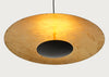 A modern pendant light with a large, shallow circular shade made of textured, warm-toned paper or resin, featuring a central black circular cutout and a glowing warm yellow light from within. The lamp is suspended by a thin black cord against a solid, light gray background.