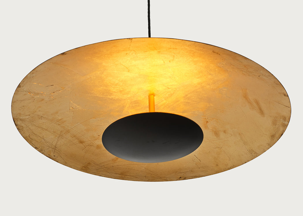 A modern pendant light with a large, shallow circular shade made of textured, warm-toned paper or resin, featuring a central black circular cutout and a glowing warm yellow light from within. The lamp is suspended by a thin black cord against a solid, light gray background.