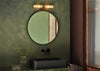 A round wall mirror with a dark frame hangs on a textured olive green bathroom wall above a black rectangular vessel sink. A modern wall-mounted light fixture is positioned above the mirror, and a glass vase with dried stems sits on the counter below.