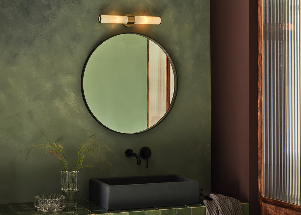A round wall mirror with a dark frame hangs on a textured olive green bathroom wall above a black rectangular vessel sink. A modern wall-mounted light fixture is positioned above the mirror, and a glass vase with dried stems sits on the counter below.