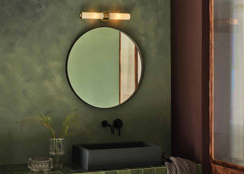 A round wall mirror with a dark frame hangs on a textured olive green bathroom wall above a black rectangular vessel sink. A modern wall-mounted light fixture is positioned above the mirror, and a glass vase with dried stems sits on the counter below.