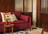 A red upholstered sofa in a warm, wood-paneled living room setting, styled with patterned throw pillows, a small wooden side table, and a floor lamp.