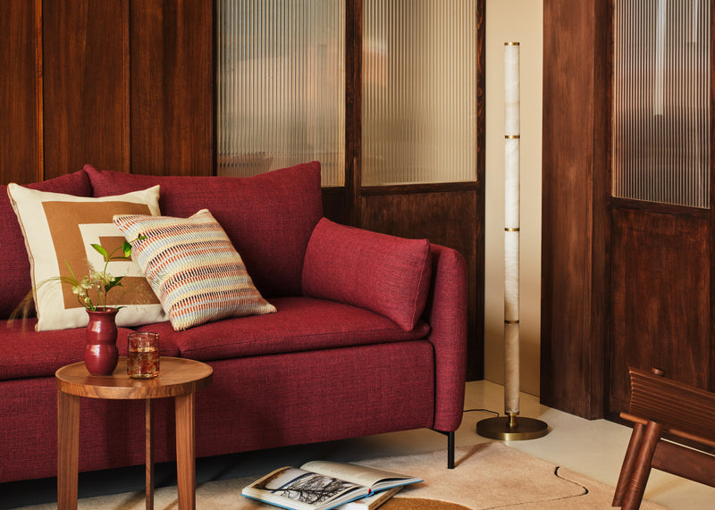 A red upholstered sofa in a warm, wood-paneled living room setting, styled with patterned throw pillows, a small wooden side table, and a floor lamp.