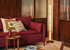 A modern red fabric sofa in a warm, wood-paneled living room setting, styled with patterned throw pillows, a small wooden side table, and a tall glowing floor lamp.
