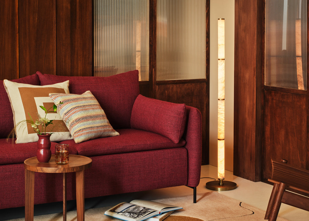 A modern red fabric sofa in a warm, wood-paneled living room setting, styled with patterned throw pillows, a small wooden side table, and a tall glowing floor lamp.