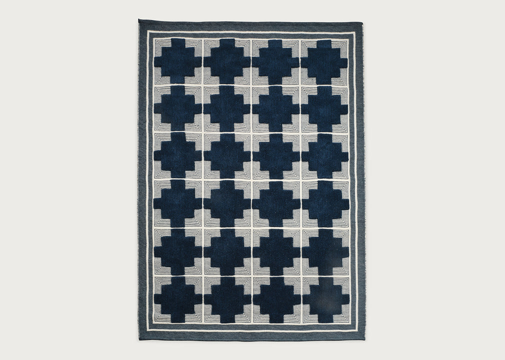 A rectangular area rug with a geometric pattern of dark navy blue crosses arranged in a grid over a light beige background, finished with a dark navy border.