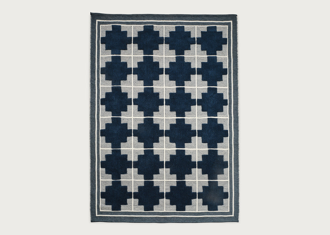 A rectangular area rug with a geometric pattern of dark navy blue crosses arranged in a grid over a light beige background, finished with a dark navy border.