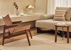 A mid-century modern wooden armchair with woven slatted back and seat, styled in a warm, minimalist living room next to a beige sofa and a small wooden side table.