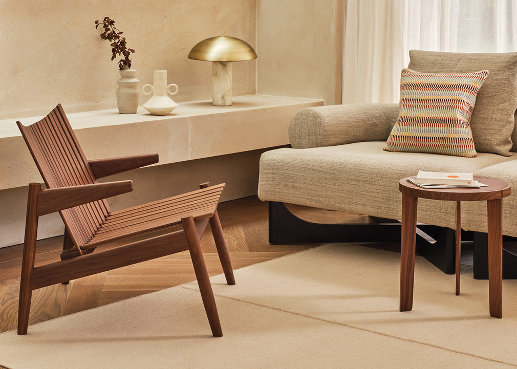 A mid-century modern wooden armchair with woven slatted back and seat, styled in a warm, minimalist living room next to a beige sofa and a small wooden side table.