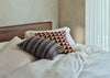 A styled bedroom scene showing a bed with cream and beige striped bedding, a dark wood headboard, and two decorative pillows. One pillow features a geometric pattern in burgundy and beige, and the other has a striped pattern in brown, beige, and white.