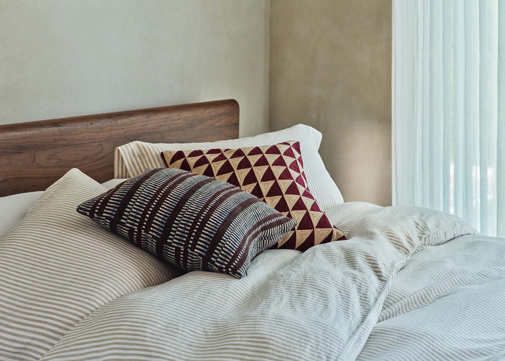 A styled bedroom scene showing a bed with cream and beige striped bedding, a dark wood headboard, and two decorative pillows. One pillow features a geometric pattern in burgundy and beige, and the other has a striped pattern in brown, beige, and white.
