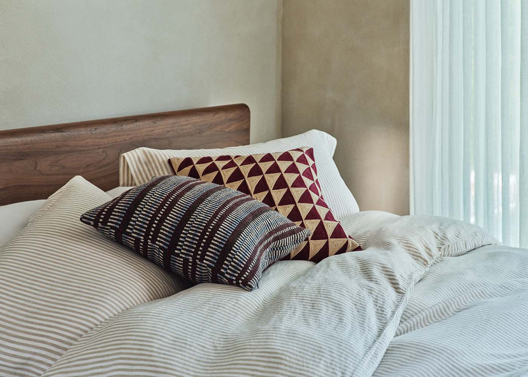 A styled bedroom scene showing a bed with cream and beige striped bedding, a dark wood headboard, and two decorative pillows. One pillow features a geometric pattern in burgundy and beige, and the other has a striped pattern in brown, beige, and white.