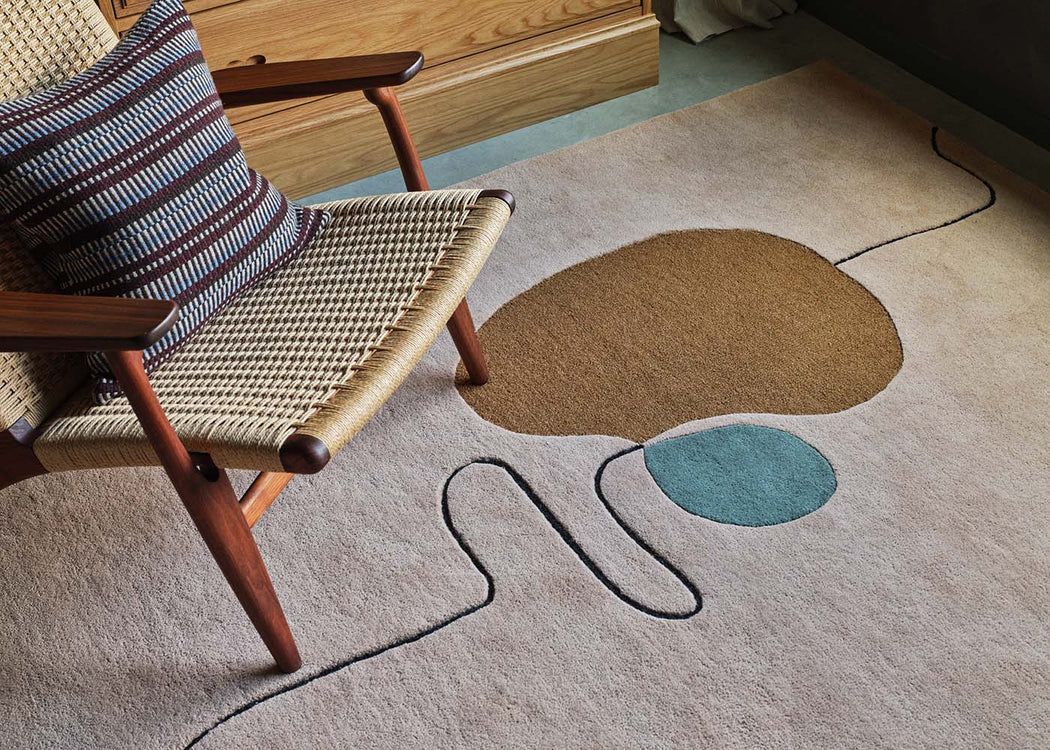 A modern abstract area rug in a styled room setting, featuring a beige base with a large brown organic shape, a teal oval, and a black wavy line. A mid-century modern woven armchair with a striped pillow is positioned on the left.