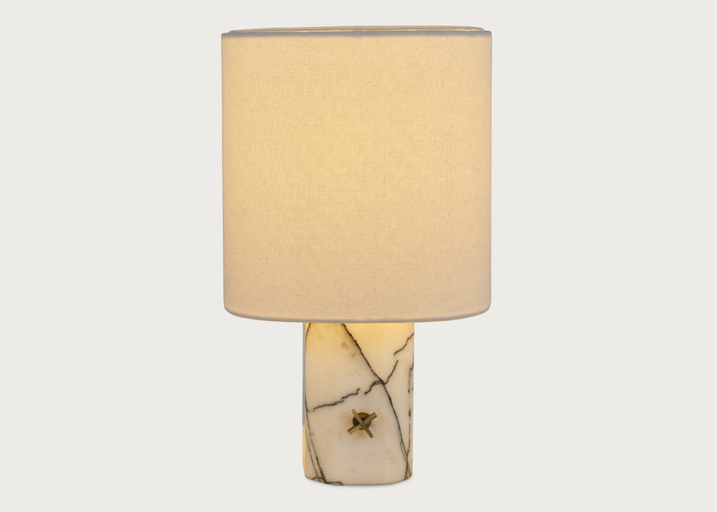 A table lamp with a cylindrical white fabric shade and a polished cream-colored stone base featuring natural dark veining and a small central brass accent, shown against a light gray background.