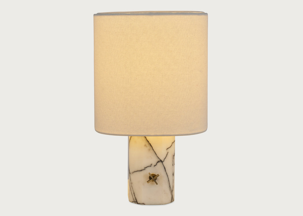 A table lamp with a cylindrical white fabric shade and a polished cream-colored stone base featuring natural dark veining and a small central brass accent, shown against a light gray background.