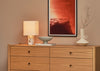 A warm wood dresser in a styled room setting, decorated with a lit table lamp, a gray bowl on a book, a white vase with dried branches, and a framed red-toned artwork on the wall above.