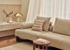 A beige upholstered sectional sofa in a warm, modern living room setting, styled with neutral throw pillows and a striped accent pillow, next to a small wooden side table.