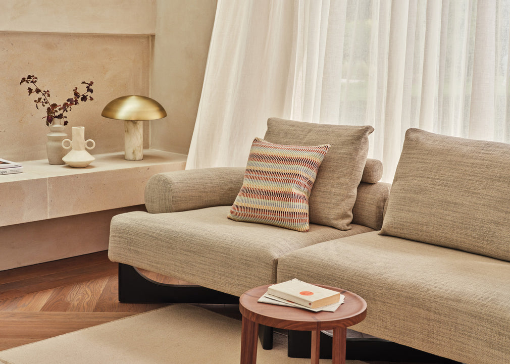 A beige upholstered sectional sofa in a warm, modern living room setting, styled with neutral throw pillows and a striped accent pillow, next to a small wooden side table.