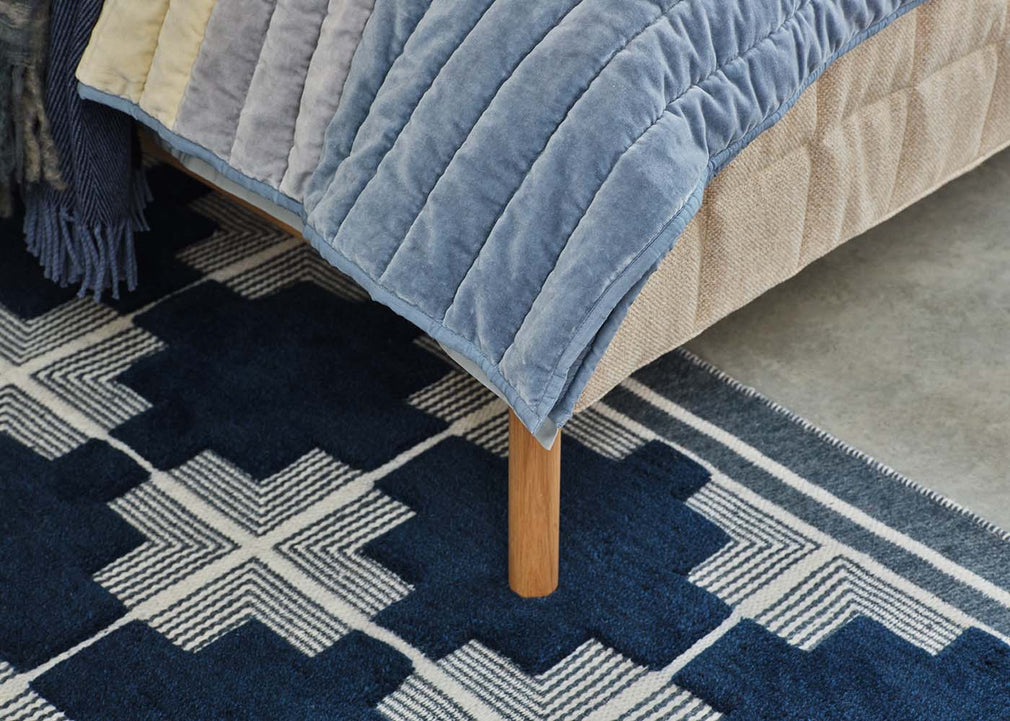 A close-up of a bed corner with a blue quilted velvet throw blanket draped over a beige upholstered frame, standing on a dark blue and cream geometric patterned rug.