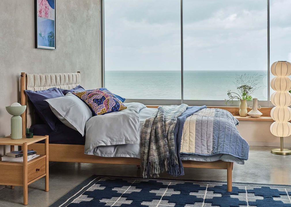 A modern wooden bed frame styled with blue, gray, and patterned bedding, set in a contemporary bedroom with a large window overlooking the ocean.