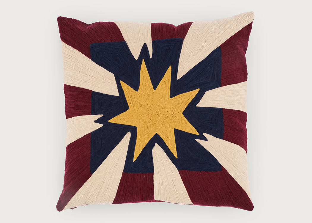 A square decorative throw pillow featuring a stylized starburst design with a central yellow star, navy blue center, and alternating cream and burgundy rays, shown against a plain light gray background.