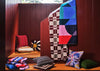 A styled interior scene with multiple colorful textiles and pillows arranged on wooden benches against dark red paneled walls. A large folded textile with bold geometric blocks in blue, pink, red, black, and white is the most prominent item, with additional patterned cushions and folded blankets around it.