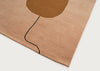 A close-up of a plush area rug with a minimalist design, featuring a dusty pink background, a large brown abstract shape near the top, and a thin black curved line running down the center, shown on a light gray background.