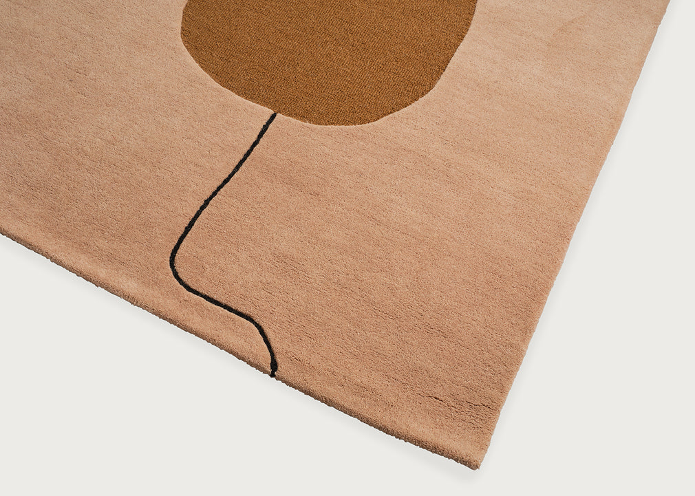 A close-up of a plush area rug with a minimalist design, featuring a dusty pink background, a large brown abstract shape near the top, and a thin black curved line running down the center, shown on a light gray background.