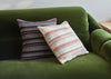 Two decorative throw pillows with striped patterns resting on a green velvet sofa. One pillow has dark brown, white, and light blue stripes, and the other has orange, white, and light blue stripes.
