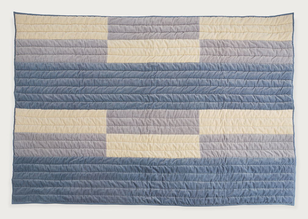 A rectangular quilted blanket with a geometric striped pattern in shades of blue, gray, and cream, displayed flat against a plain white background.