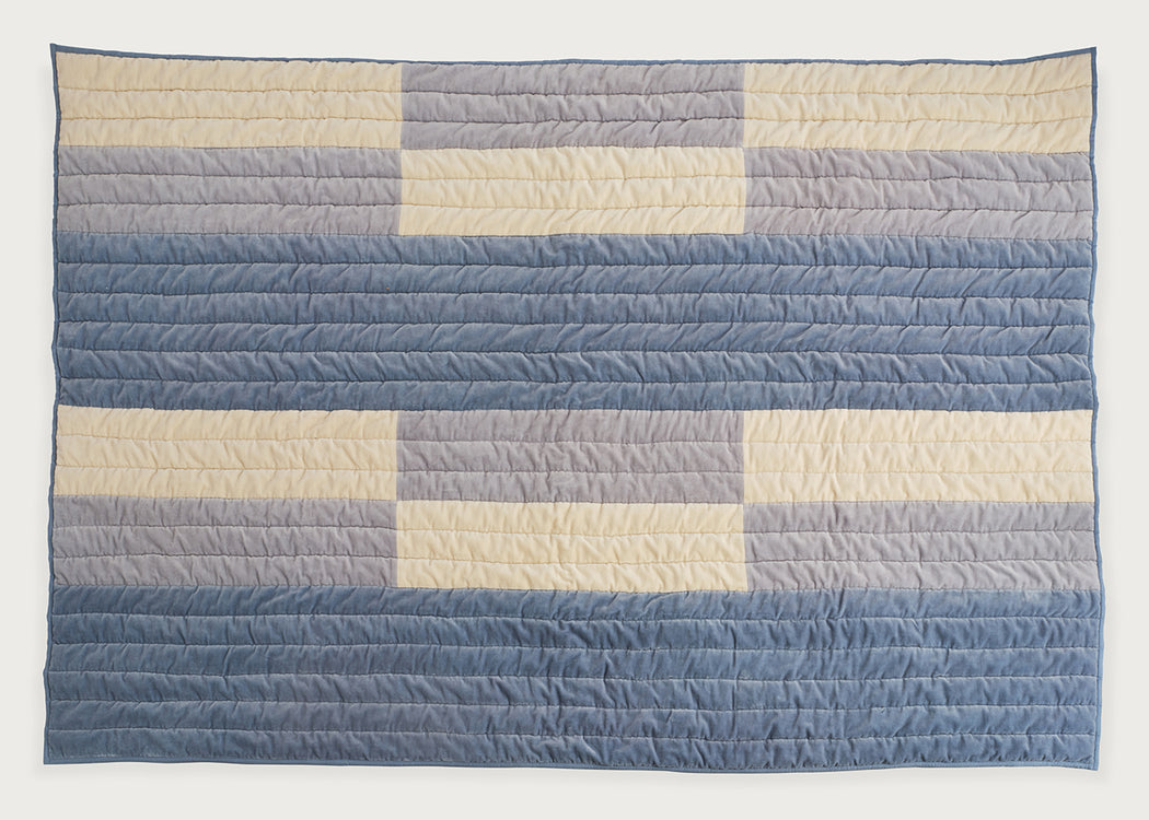 A rectangular quilted blanket with a geometric striped pattern in shades of blue, gray, and cream, displayed flat against a plain white background.