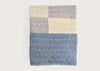 A quilted blanket with a geometric color-blocked pattern featuring a large dusty blue section, a gray section, and cream sections, shown flat against a light off-white background.