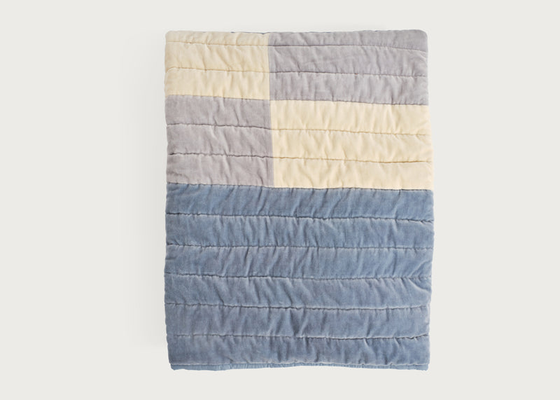 A quilted blanket with a geometric color-blocked pattern featuring a large dusty blue section, a gray section, and cream sections, shown flat against a light off-white background.